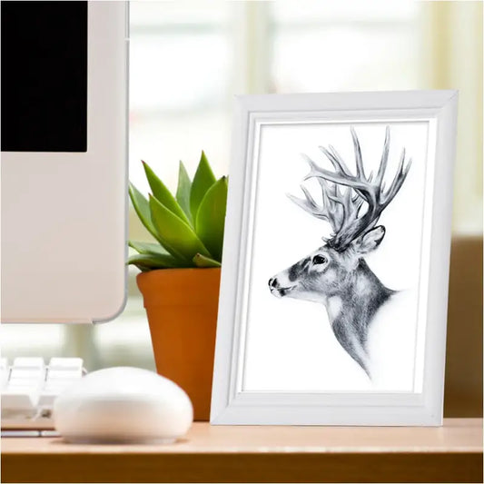 Een zwart-wit tekening van een hert met imposante geweien, ingelijst in een witte lijst, staat op een bureau naast een potted succulent en een computer. Perfect voor een minimalistische kantoor- of woonruimte.