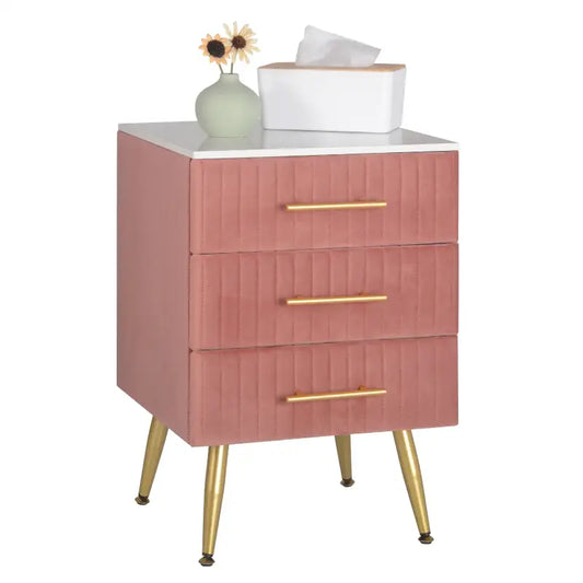 Een roze, gestoffeerde nachtkastje met drie laden en goudkleurige handgrepen, voorzien van elegante, goudkleurige poten. Ideaal voor een moderne slaapkamer of als nachtkastje in een stijlvolle woonruimte.