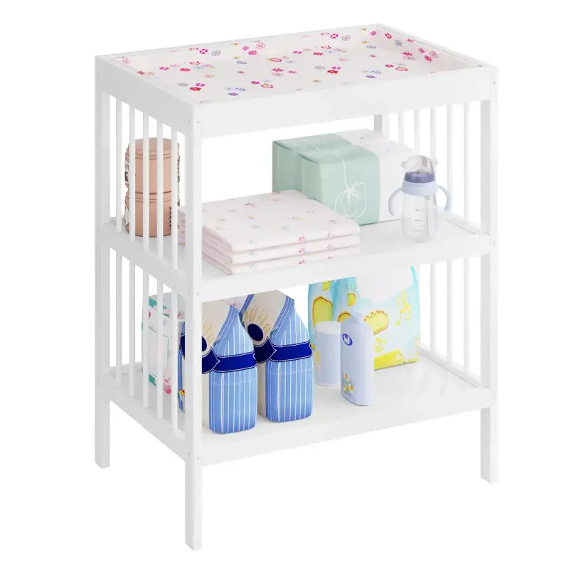 Een witte babyverzorgingskast met drie planken, bedekt met een kleurrijk bloemenpatroon. Voorzien van babyartikelen zoals luiers, flesjes en washandjes voor babyverzorging.