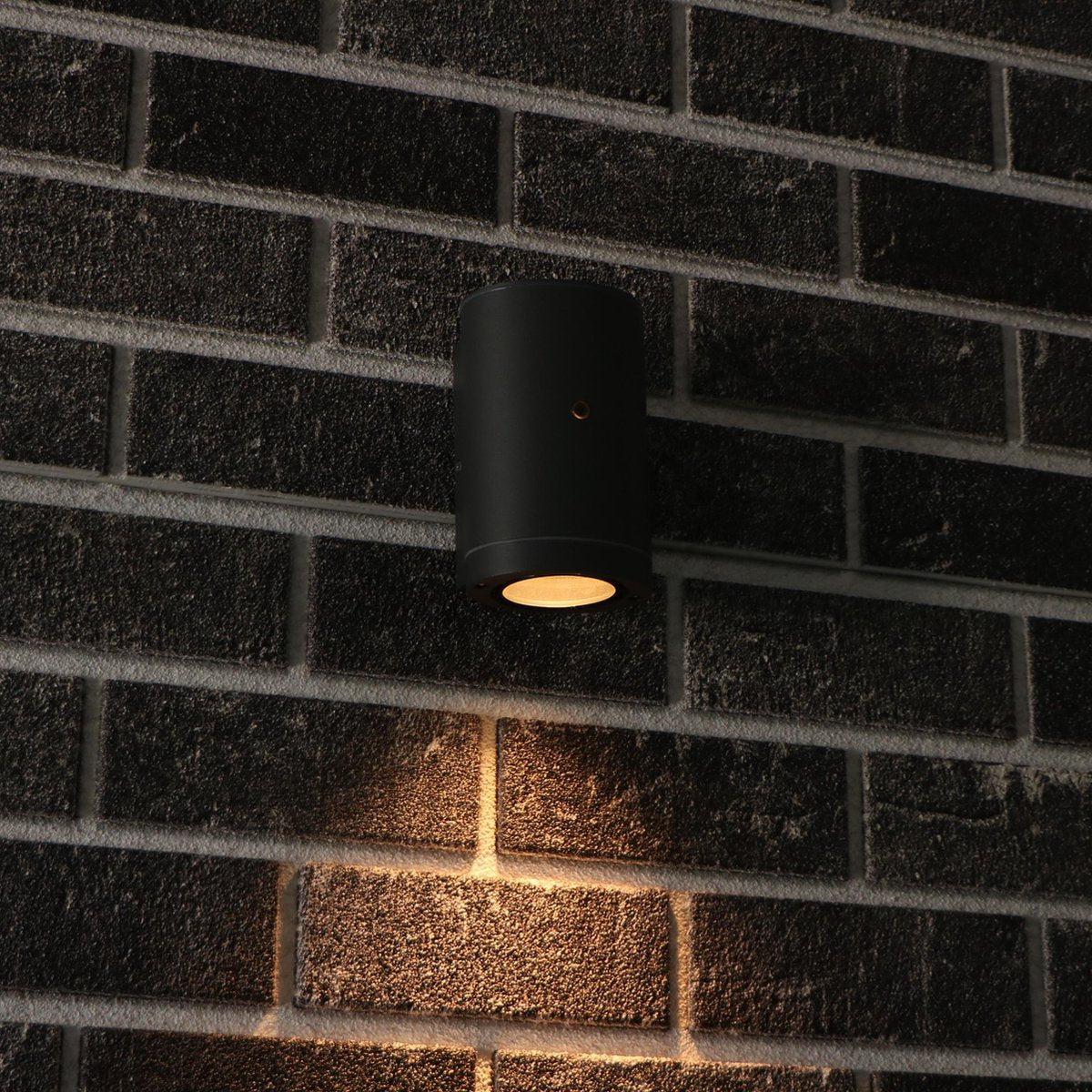 Zwart cilindrische wandlamp met goudkleurige rand en warm wit licht, geschikt voor moderne interieurs. Compacte design met zichtbare montagegaten voor wandmontage.
