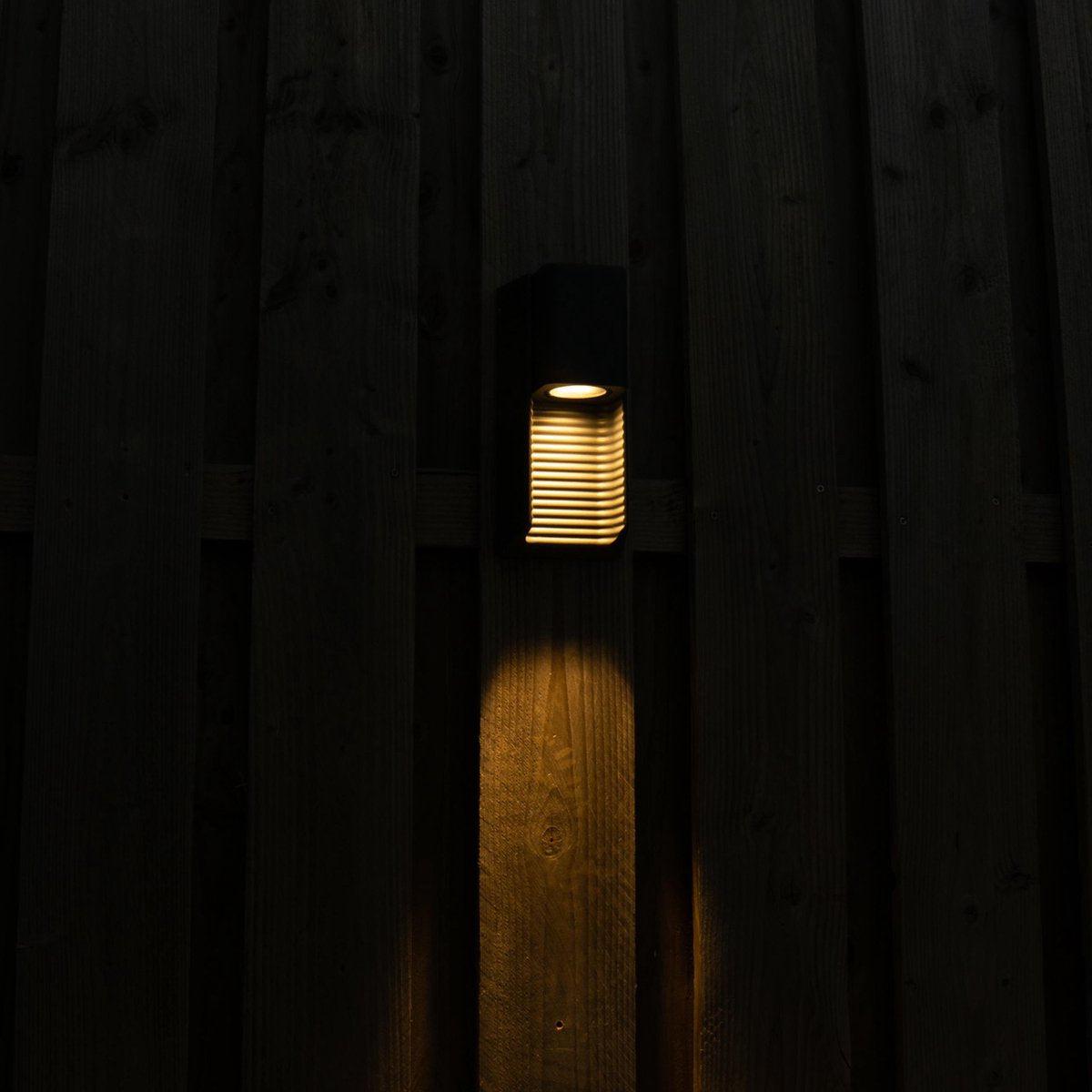 Zwart buitenlamp met horizontale lamellen, die warm licht werpt op een houten schutting. Ideaal voor tuinverlichting en nachtelijke sfeer in de buitenruimte.