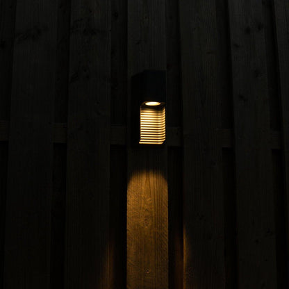 Zwart buitenlamp met horizontale lamellen, die warm licht werpt op een houten schutting. Ideaal voor tuinverlichting en nachtelijke sfeer in de buitenruimte.