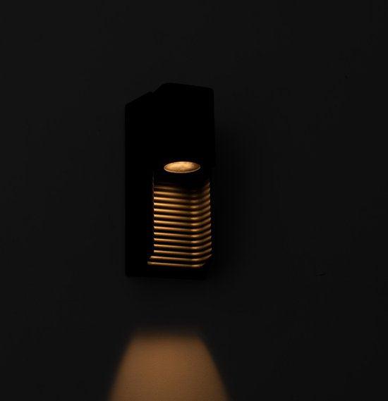 Zwart wandlamp met horizontale lamellen, die warm licht werpt op een houten schutting. Ideaal voor buitenverlichting en tuinverlichting, met een moderne, minimalistische uitstraling.