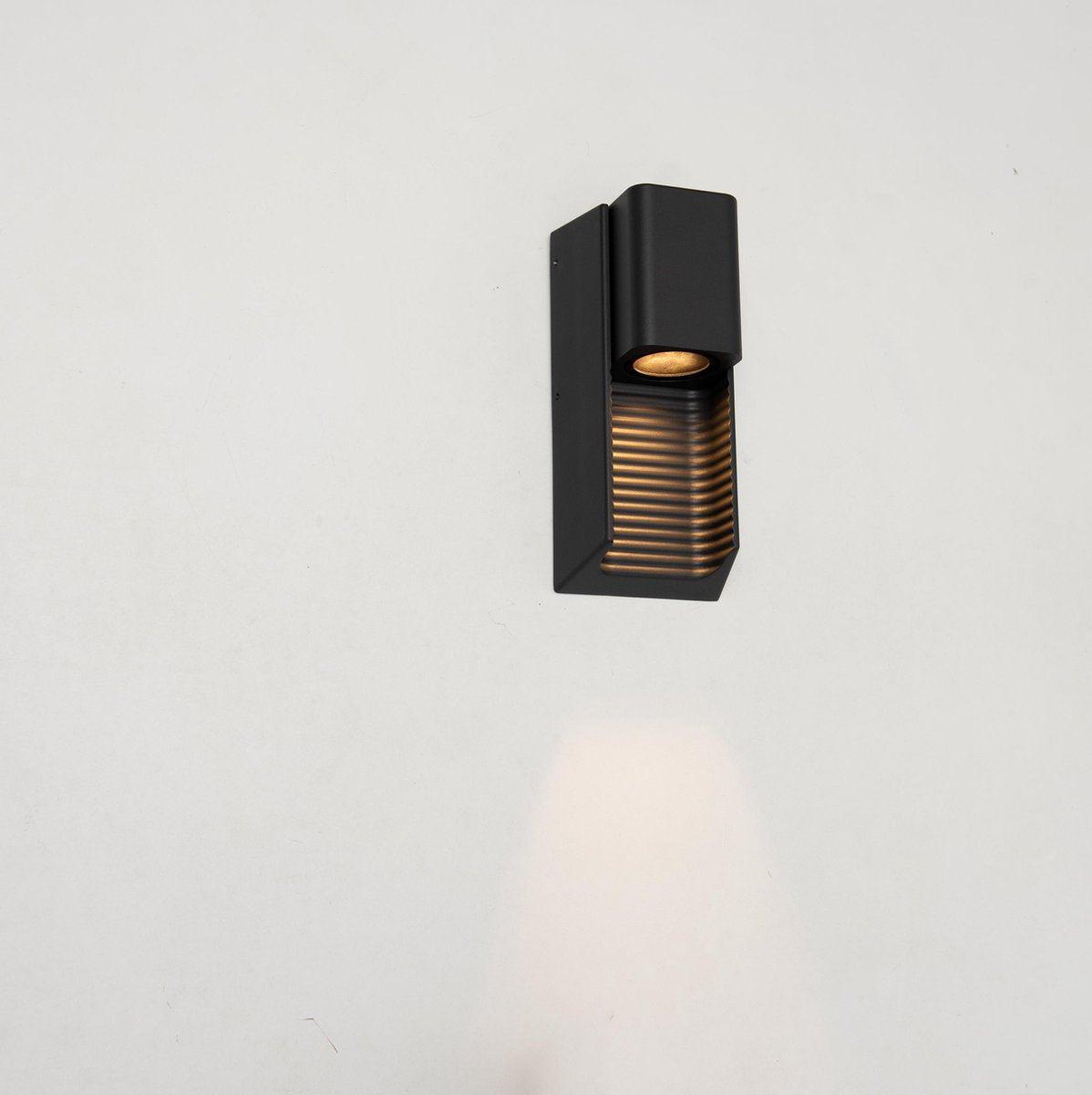 Zwart buitenlamp met horizontale lamellen, die warm licht werpt op een houten schutting. Ideaal voor tuinverlichting en nachtelijke sfeercreatie.