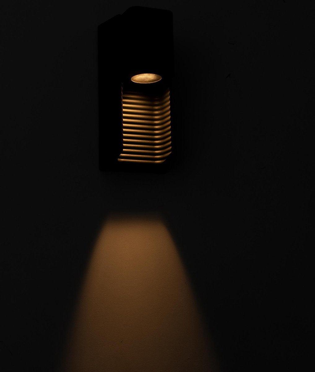 Zwarte wandlamp met horizontale lamellen, die warm licht uitstraalt op een houten schutting. Ideaal voor buitenverlichting en tuinverlichting, met een moderne, minimalistische uitstraling.