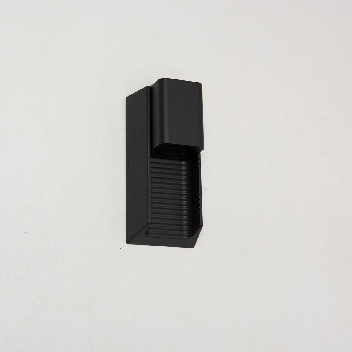 Zwart wandlamp met horizontale lamellen, die warm licht verspreidt op een houten schutting. Ideaal voor buitenverlichting en tuinverlichting, met een moderne, minimalistische uitstraling.