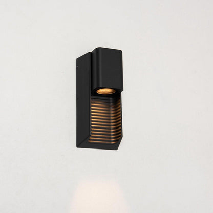 Zwart buitenlamp met horizontale lamellen, die warm wit licht uitstraalt op een houten schutting. Ideaal voor tuinverlichting en nachtelijke veiligheid.