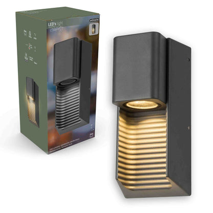 Zwart buitenlamp met horizontale lamellen, die warm licht werpt op een houten schutting. Ideaal voor tuinverlichting en nachtelijke sfeercreatie.