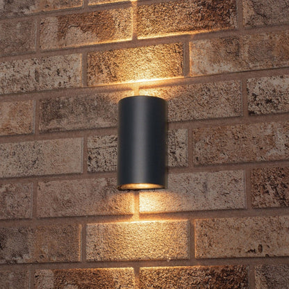 Zwart cilindrische wandlamp met warm wit licht, geïnstalleerd op een textuurrijke bakstenen muur. Ideaal voor buitenverlichting en moderne tuin- of terrasdecoratie.