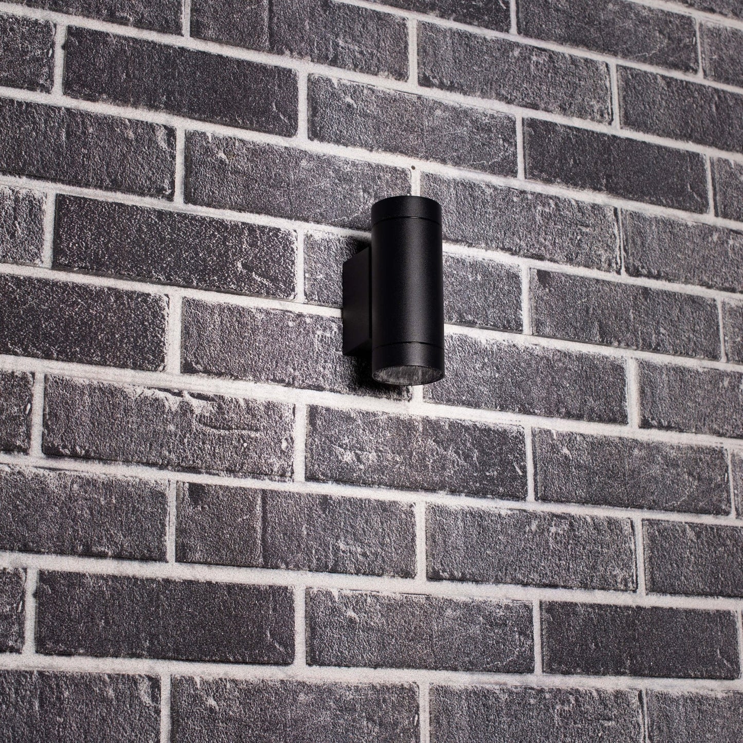 Zwart, cilindrisch buitenspot met warm wit licht, bevestigd aan een grijze bakstenen muur. Modern design voor buitenverlichting en tuinverlichting.
