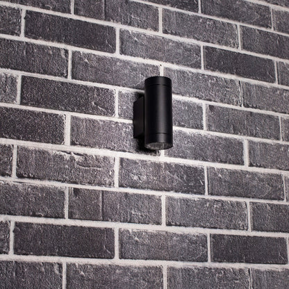 Zwart, cilindrisch buitenspot met warm wit licht, bevestigd aan een grijze bakstenen muur. Modern design voor buitenverlichting en tuinverlichting.