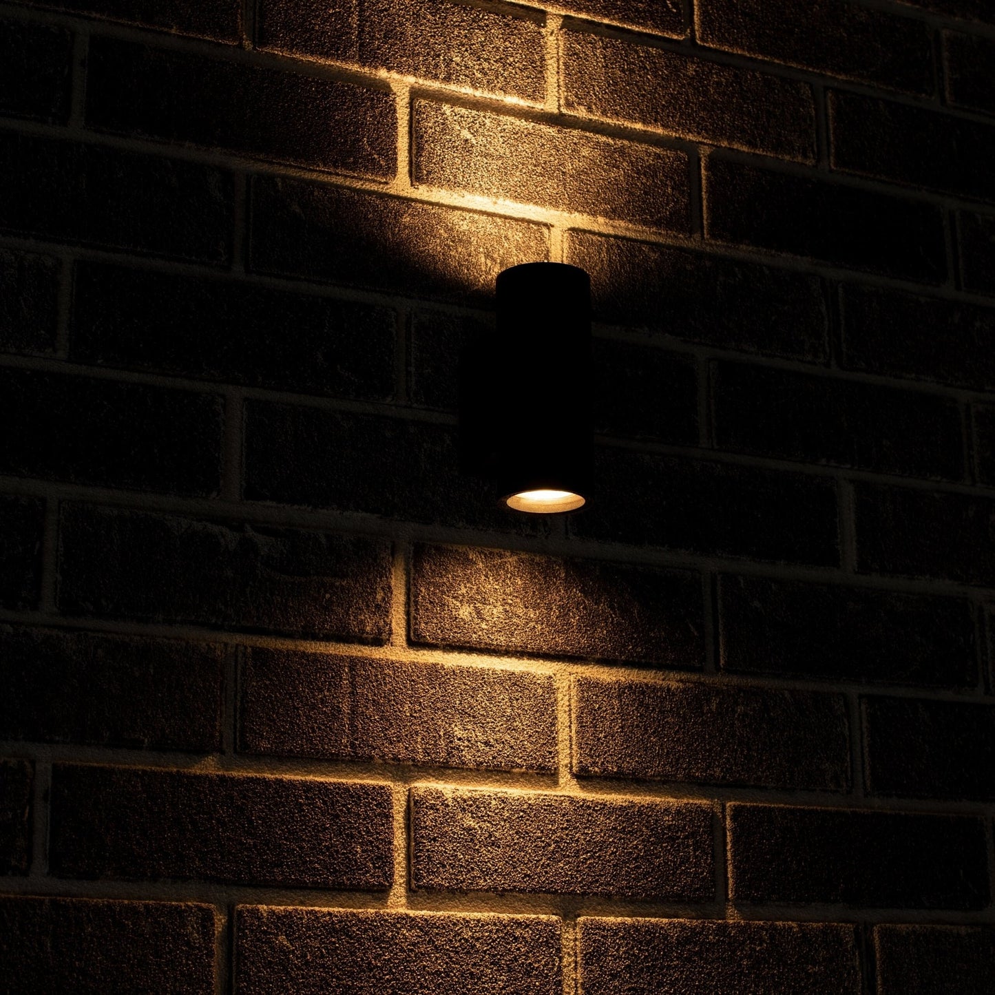 Zwart, cilindrisch buitenspot met warm wit licht, gemonteerd op een grijze bakstenen muur. Modern design voor buitenverlichting en tuinverlichting.