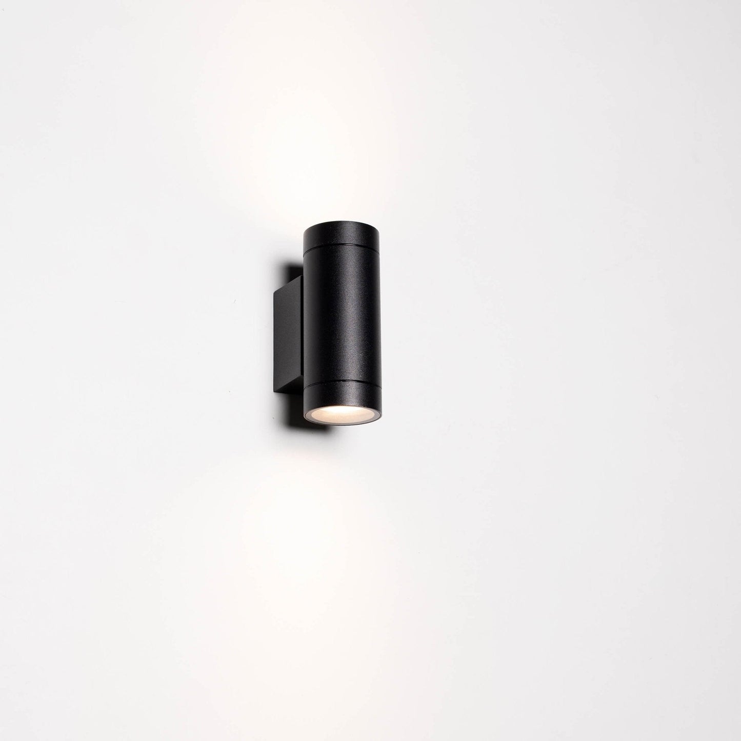 Zwart, cilindrisch buitenspot met warm wit licht, gemonteerd op een grijze bakstenen muur. Modern design voor buitenverlichting en tuinverlichting.