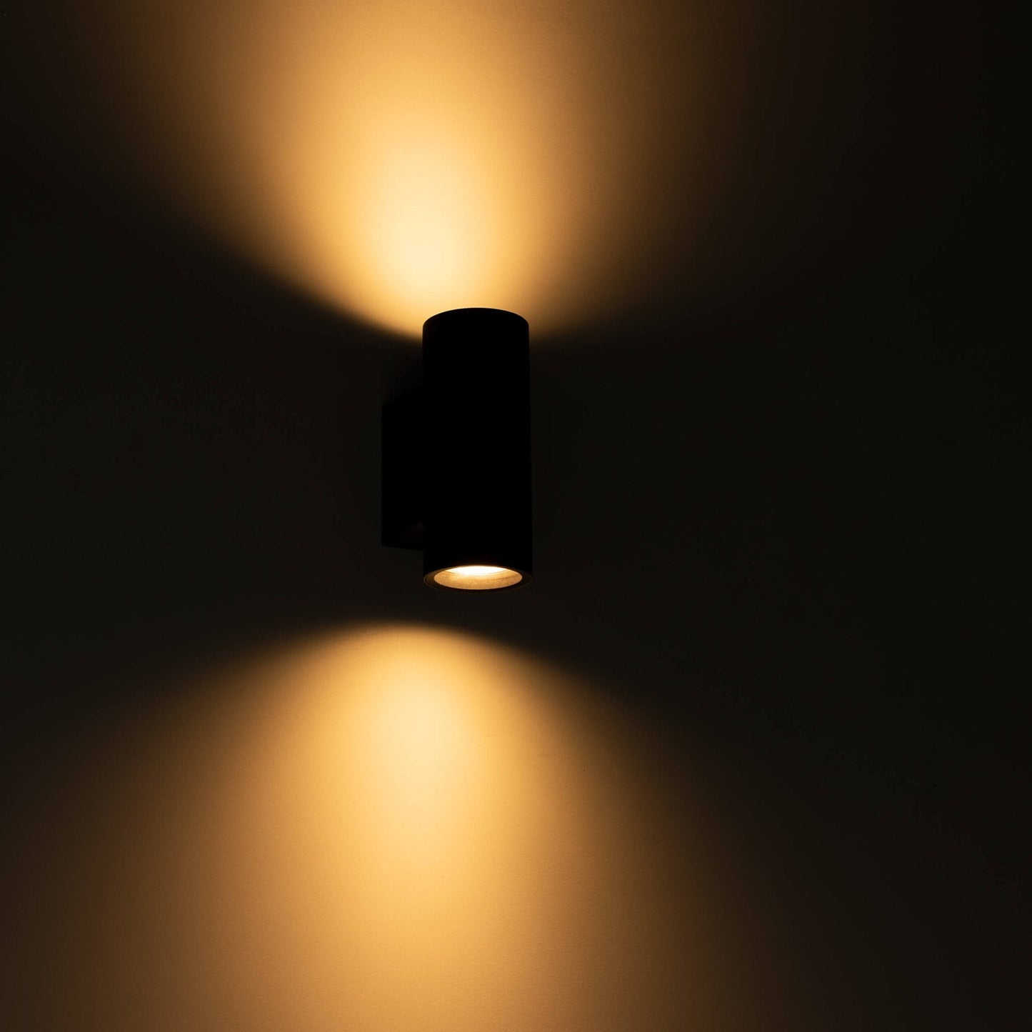 Zwart, cilindrisch buitenspot met warm wit licht, bevestigd aan een grijze bakstenen muur. Modern design voor buitenverlichting en tuinverlichting.