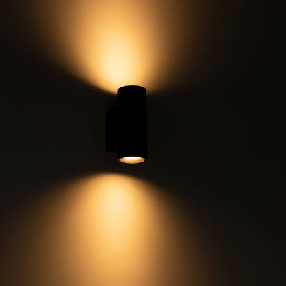 Zwart, cilindrisch buitenspot met warm wit licht, bevestigd aan een grijze bakstenen muur. Modern design voor buitenverlichting en tuinverlichting.