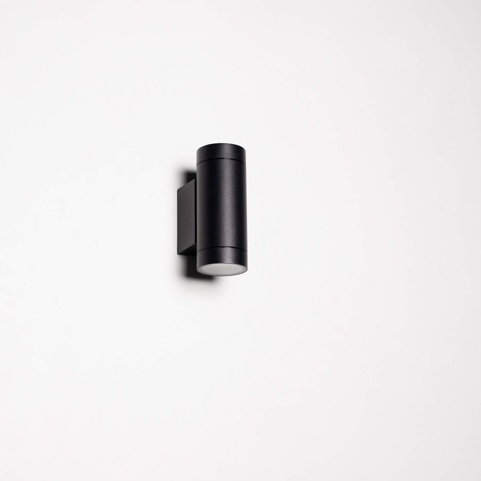 Zwart, cilindrisch buitenspot met warm wit licht, gemonteerd op een grijze bakstenen muur. Modern design voor buitenverlichting en tuinverlichting.