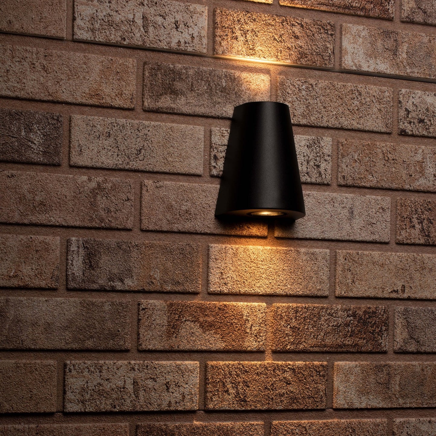 Zwart, cilindrisch wandlampje met warm wit licht, bevestigd aan een textuurrijke betonmuur. Perfect voor moderne buitenverlichting en architectonische accentverlichting.