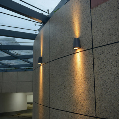 Zwart, cilindrisch wandlampje met warm wit licht, bevestigd aan een textuurrijke betonmuur. Ideaal voor moderne buitenverlichting en architectonische accentverlichting.