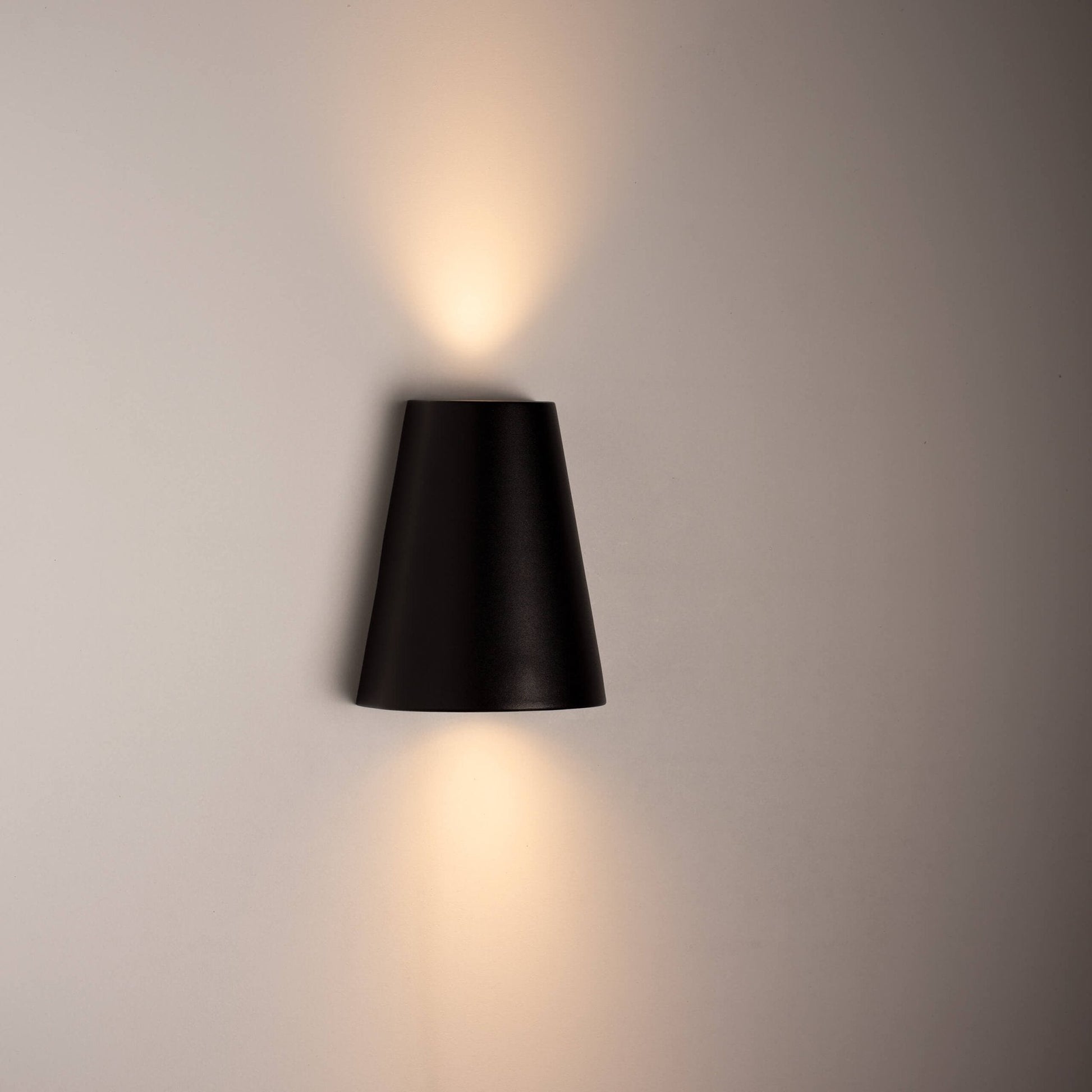 Zwart, cilindrisch wandlampje met warm wit licht, bevestigd aan een textuurrijke betonmuur. Perfect voor moderne buitenverlichting en architectonische accentverlichting.