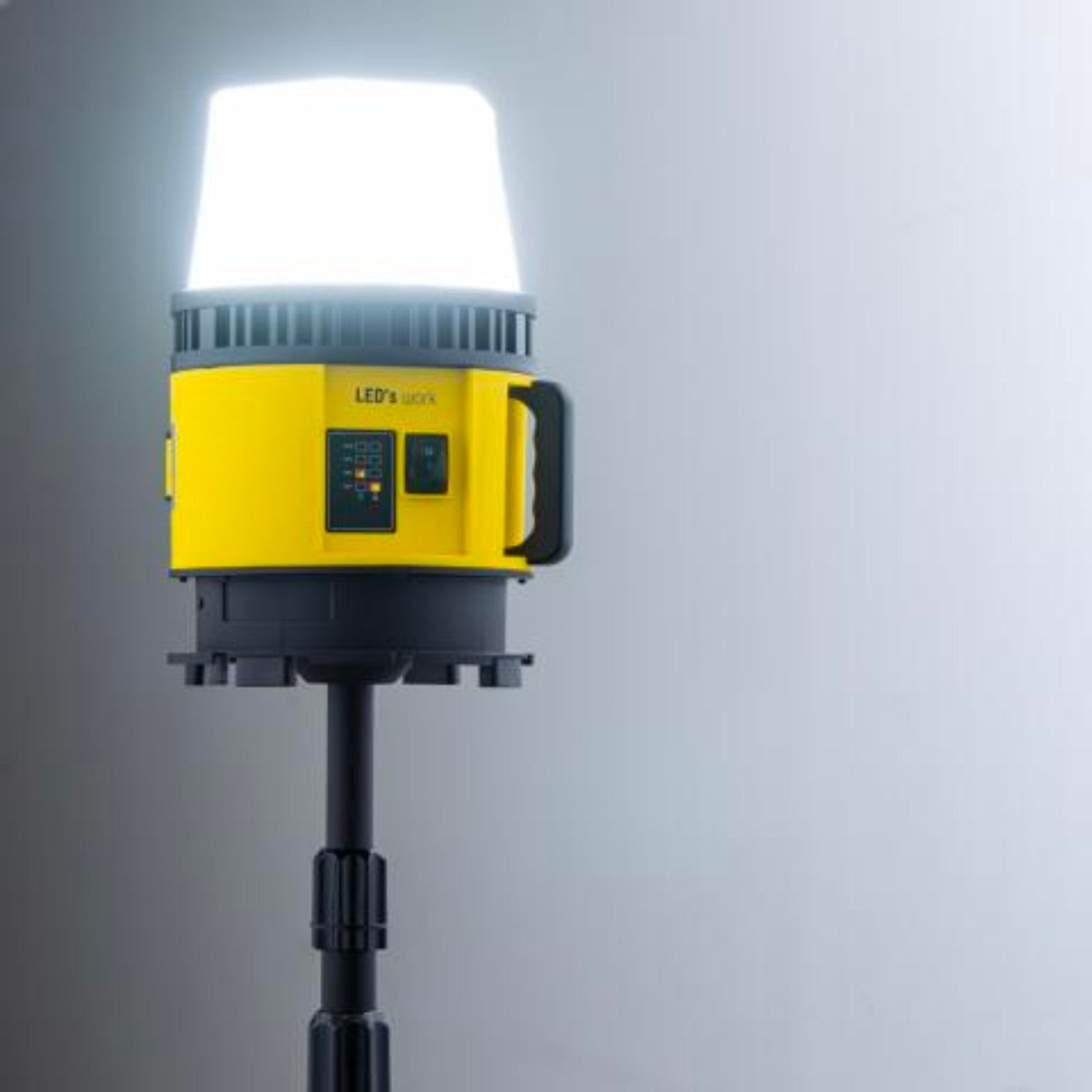 Een gele en zwarte batterijoplaadbare werklamp met witte kap en rode accu-connector, geschikt voor gebruik in bouw- en onderhoudsprojecten.