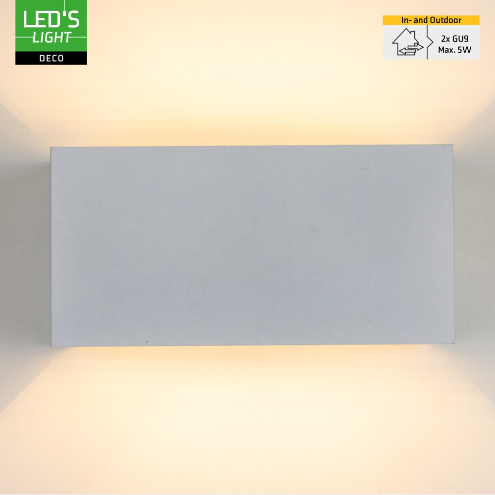 Een witte, rechthoekige wandlamp met warm wit licht, bevestigd aan een grijze bakstenen muur. Ideaal voor buitenverlichting en moderne tuin- of terrasdecoratie.