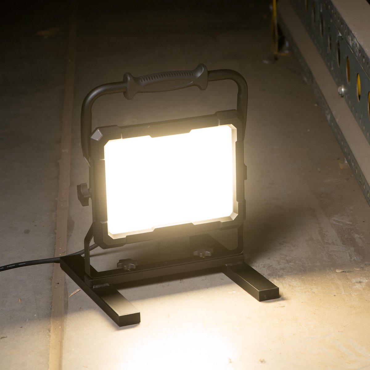 Zwart LED-verlichtingsapparaat met verstelbare arm en handgreep, voorzien van een sterk lichtpaneel en een kabel met stekker. Geschikt voor werken in donkere ruimtes of als noodverlichting.