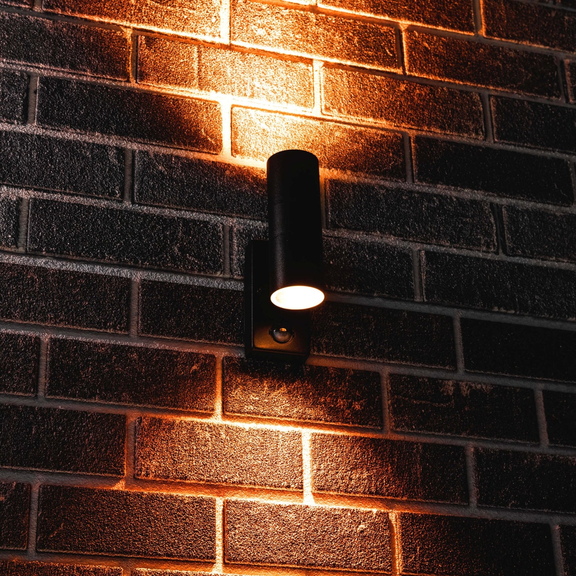Zwarte wandlamp met warm wit licht, geïnstalleerd op een donkere bakstenen muur. Modern design voor buitenverlichting, ideaal voor tuin of terras.