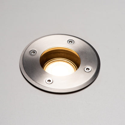 Zilverkleurige, ronde bodemplaatslamp met glazen lens en zichtbare schroeven, voorzien van een zwarte cilindrische basis. Geschikt voor tuinverlichting en buitenverlichting.