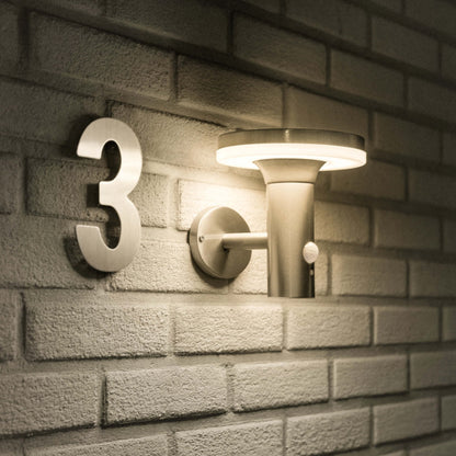 Een moderne, zonnepanelen-gevoerde wandlamp met warm wit licht, in mat zwart metaal, bevestigd aan een textuurrijke bakstenen muur. Ideaal voor buitenverlichting en veiligheid in tuinen of op gangpaden.