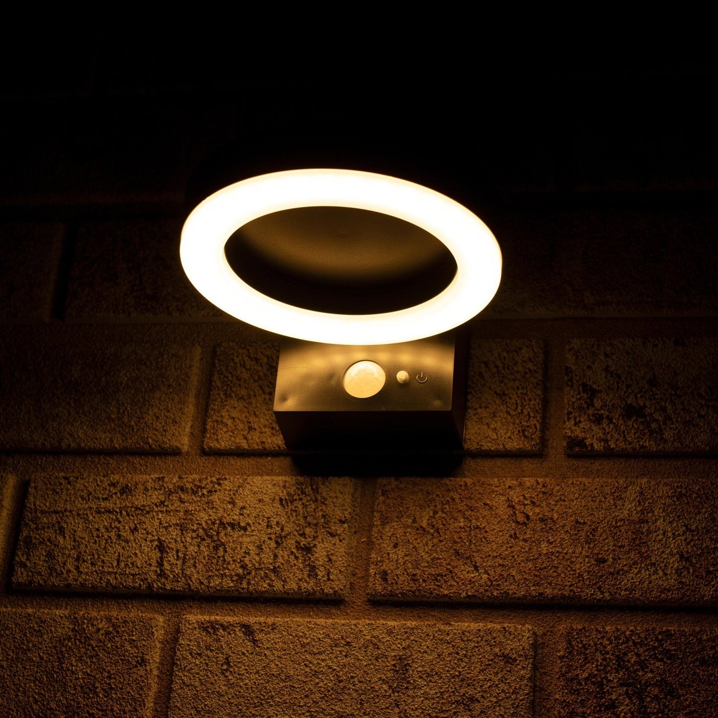Een moderne, zwarte ringvormige wandlamp met warm wit licht, gemonteerd op een textuurrijke bakstenen muur. Uitgerust met bewegingssensor en touch-knop voor buitenverlichting.