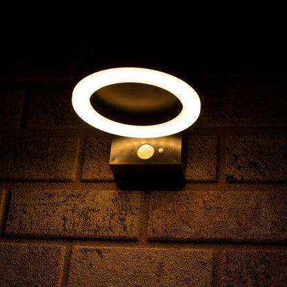 Een moderne, zwarte ringvormige wandlamp met warm wit licht, gemonteerd op een textuurrijke bakstenen muur. Uitgerust met bewegingssensor en touch-knop voor buitenverlichting.