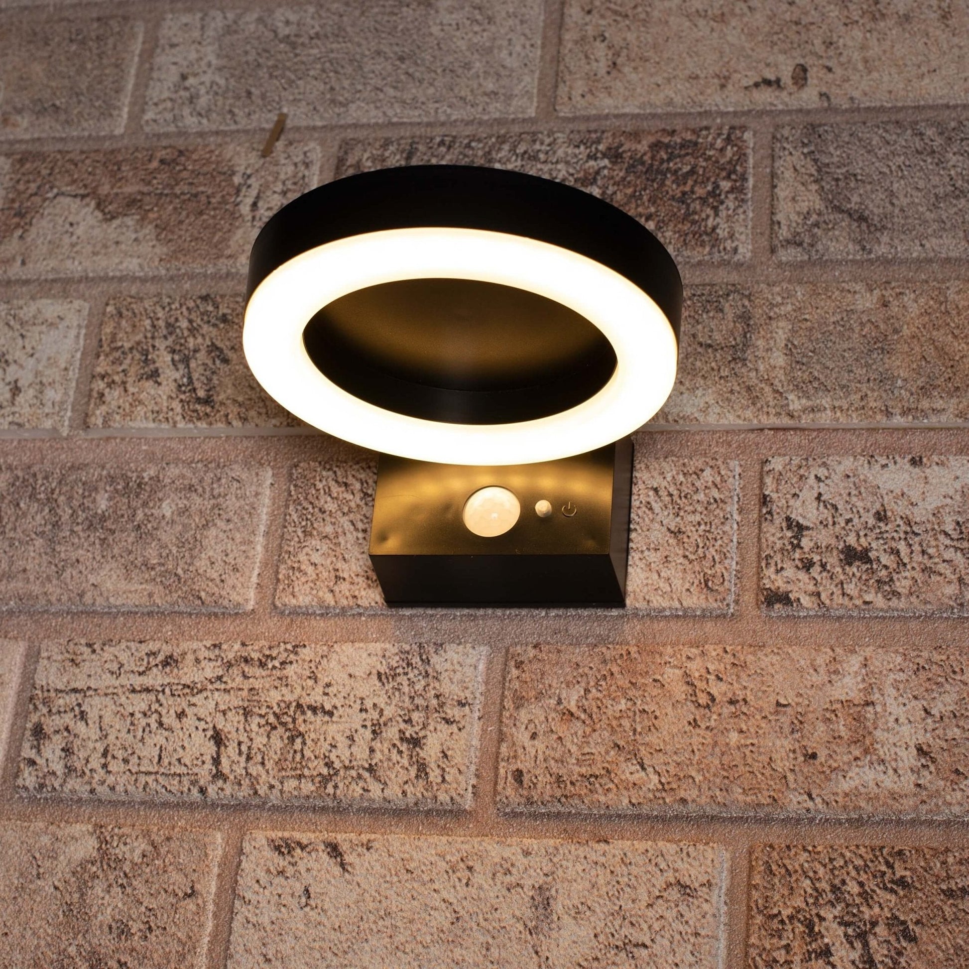 Een moderne, zwarte ringvormige wandlamp met warm wit licht, gemonteerd op een textuurrijke bakstenen muur. Uitgerust met bewegingssensor en aan/uit-knop voor gebruik als veiligheidsverlichting.