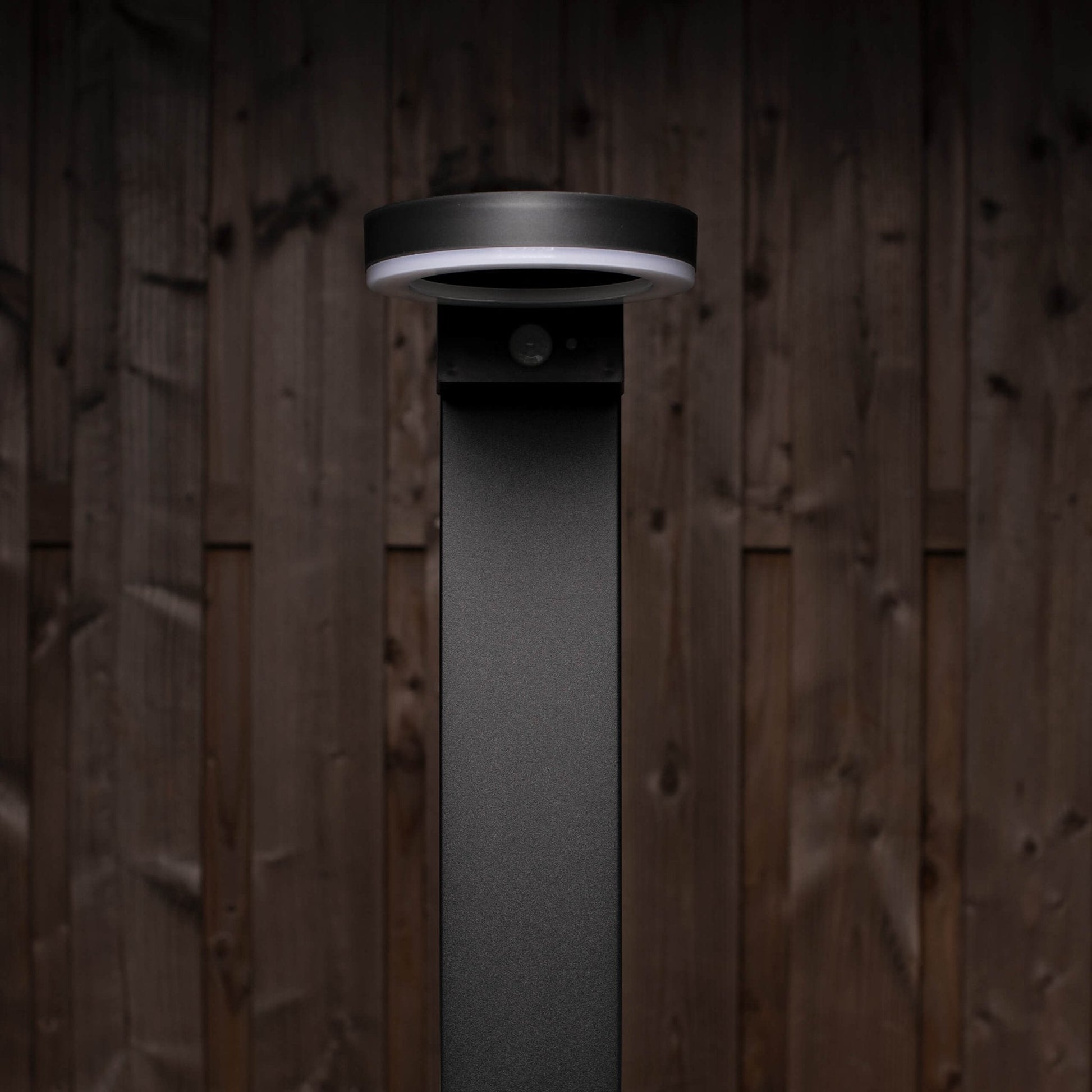 Een moderne, zwarte buitenlamp met een helder ringverlichting en een zichtbare bewegingssensor, geplaatst tegen een donkere houten achtergrond. Ideaal voor veilige verlichting in tuinen en op gangpaden.