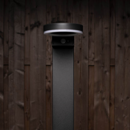 Een moderne, zwarte buitenlamp met een helder ringverlichting en een zichtbare bewegingssensor, geplaatst tegen een donkere houten achtergrond. Ideaal voor veilige verlichting in tuinen en op gangpaden.
