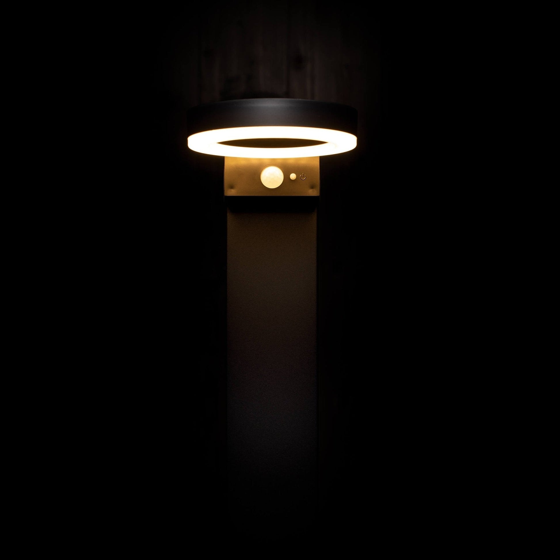 Een moderne, zwarte buitenlamp met een helder, ringvormig licht en een zichtbare bewegingssensor, geplaatst tegen een donkere houten achtergrond. Ideaal voor veilige verlichting in de tuin of op het terras.