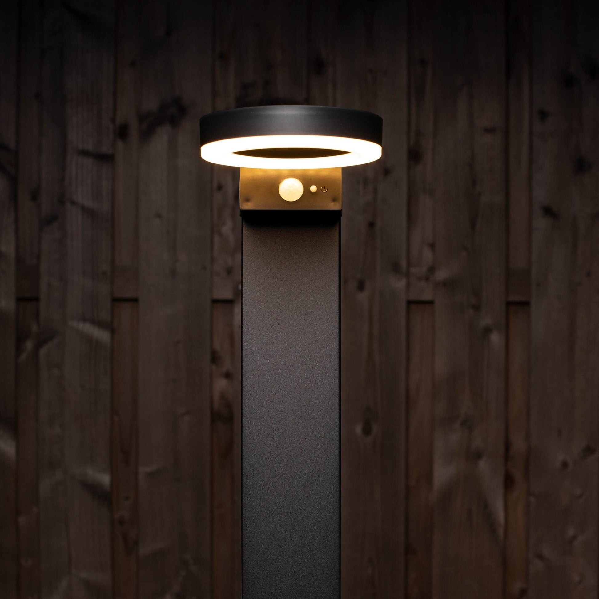 Een moderne, zwarte buitenlamp met een helder ringlicht en een zichtbare bewegingssensor, geplaatst tegen een donkere houten achtergrond. Ideaal voor veilige verlichting in tuinen en op gangpaden.