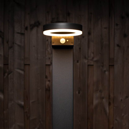 Een moderne, zwarte buitenlamp met een helder ringlicht en een zichtbare bewegingssensor, geplaatst tegen een donkere houten achtergrond. Ideaal voor veilige verlichting in tuinen en op gangpaden.