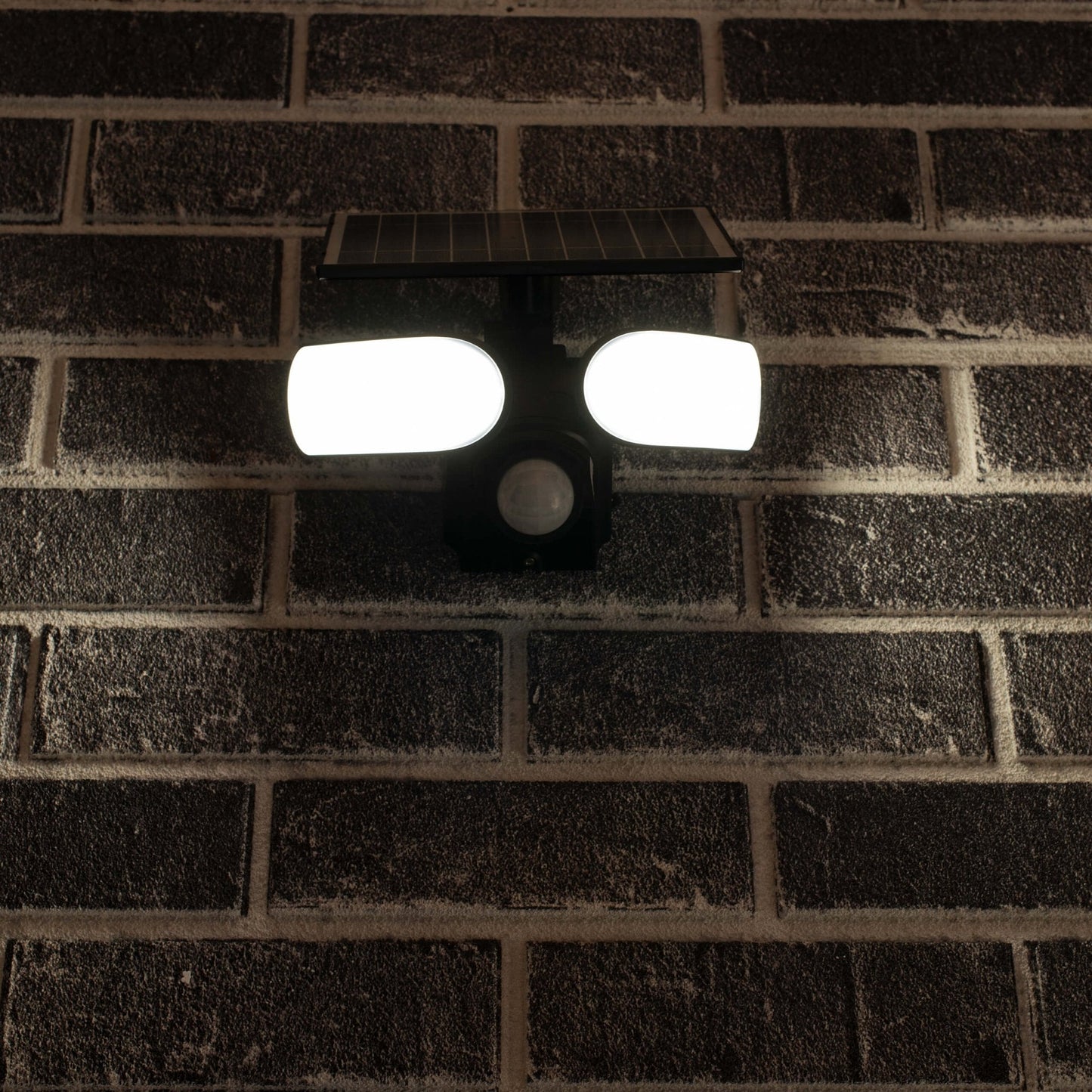 Zwarte zonnepanelenverlichting met twee heldere LED-lampen en bewegingssensor, gemonteerd op een donkere bakstenen muur. Ideaal voor buitenverlichting en nachtveiligheid.