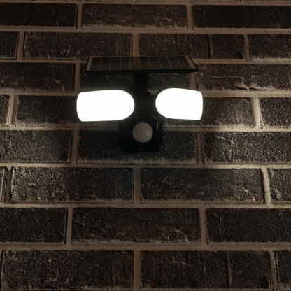 Zwarte zonnepanelenverlichting met twee heldere LED-lampen en bewegingssensor, gemonteerd op een donkere bakstenen muur. Ideaal voor buitenverlichting en nachtveiligheid.
