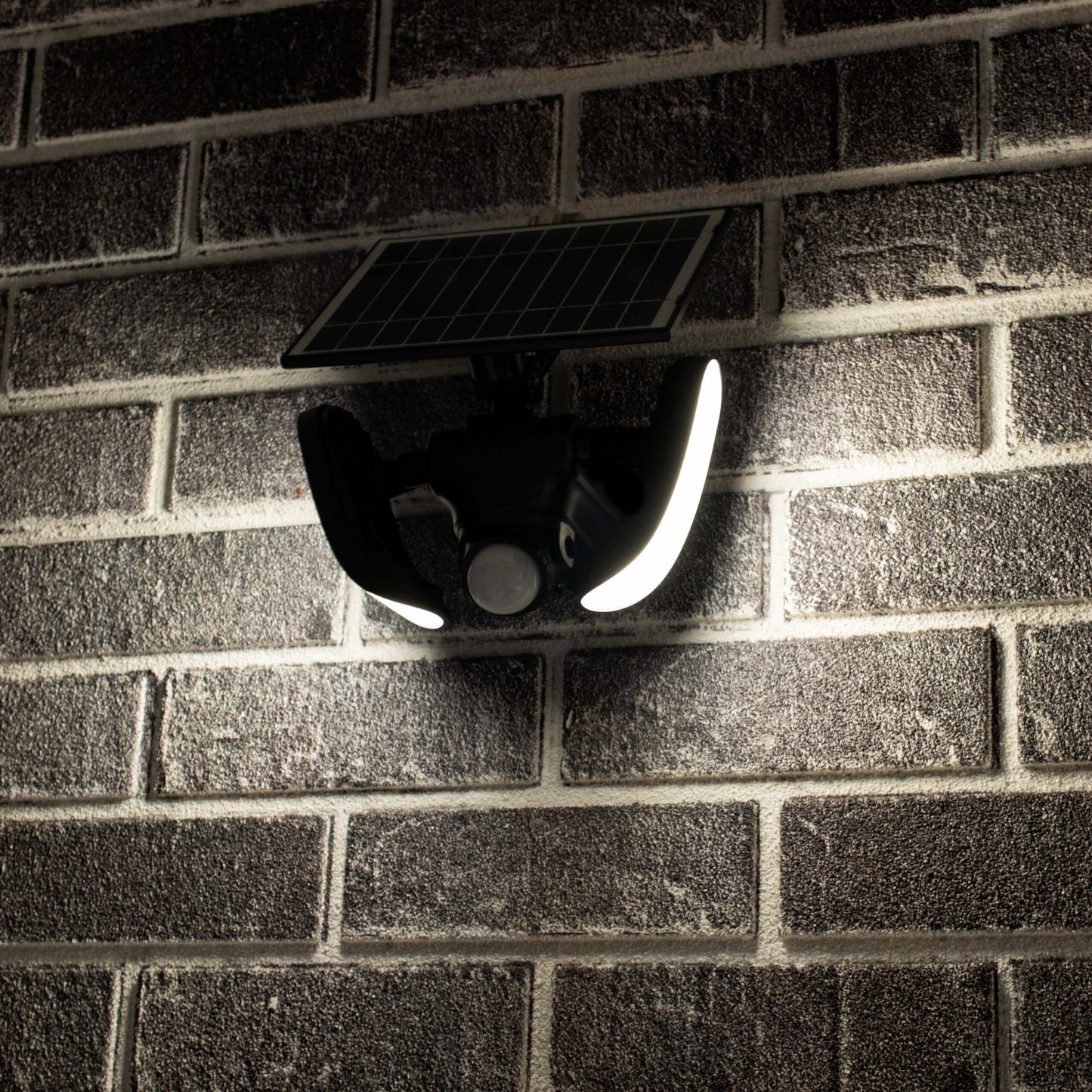 Zwarte zonnepanelenverlichting met twee heldere LED-lampen en bewegingssensor, gemonteerd op een donkere bakstenen muur. Ideaal voor veilige buitenverlichting en nachtverlichting in tuinen of op gangpaden.