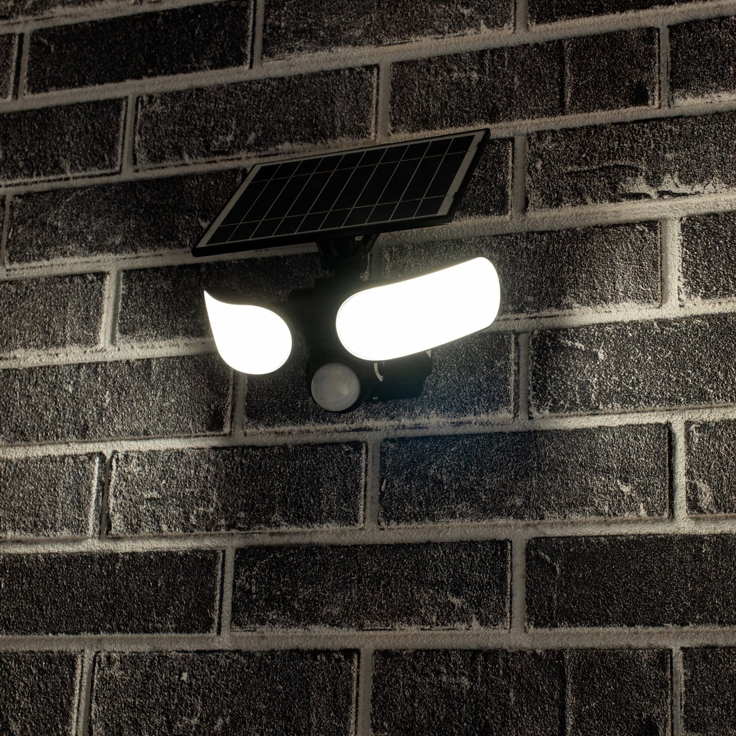 Zwarte zonnepanelenverlichting met twee heldere LED-lampen en bewegingssensor, gemonteerd op een donkere bakstenen muur. Ideaal voor nachtelijke veiligheid en energiezuinig verlichten van buitenruimtes.