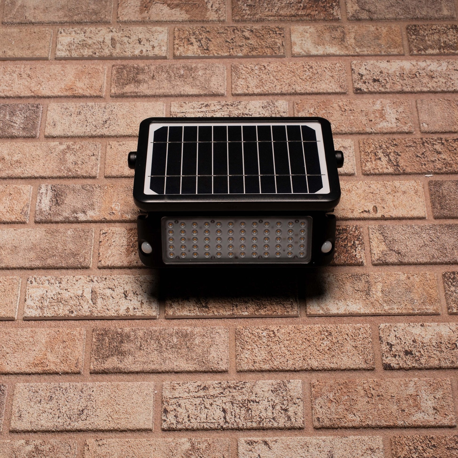 Zwarte zonnepanelenverlichting met warm wit licht, gemonteerd op een bakstenen muur. Ideaal voor buitenverlichting en tuinverlichting, met zichtbare montagebeugels en een compacte, moderne ontwerp.
