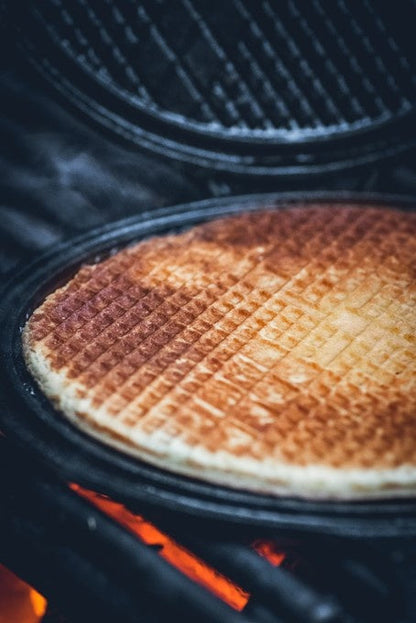Zwarte, ronde wafelpan met opengeslagen deksel en duidelijke rasterpatronen, geschikt voor het bakken van wafels of broodjes. Gemaakt van kookstalen met handgrepen voor gemakkelijke hantering.