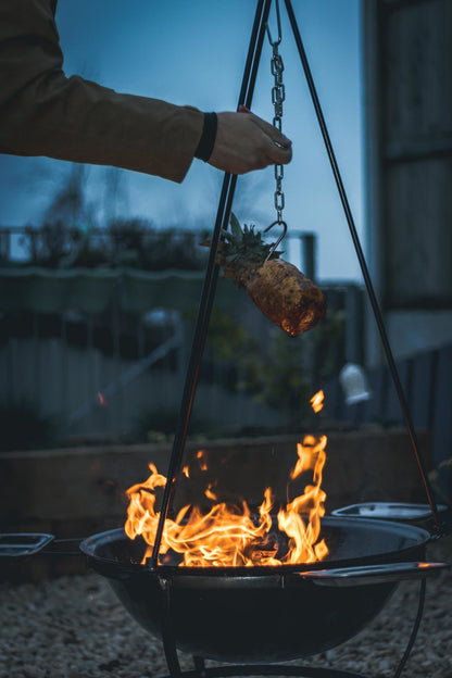 Zwarte hangende barbecue met grill en kookpan, voor buitenkeuken. Met decoratieve pootjes en metalen schepen, geschikt voor barbecuefeesten en grillavonden.