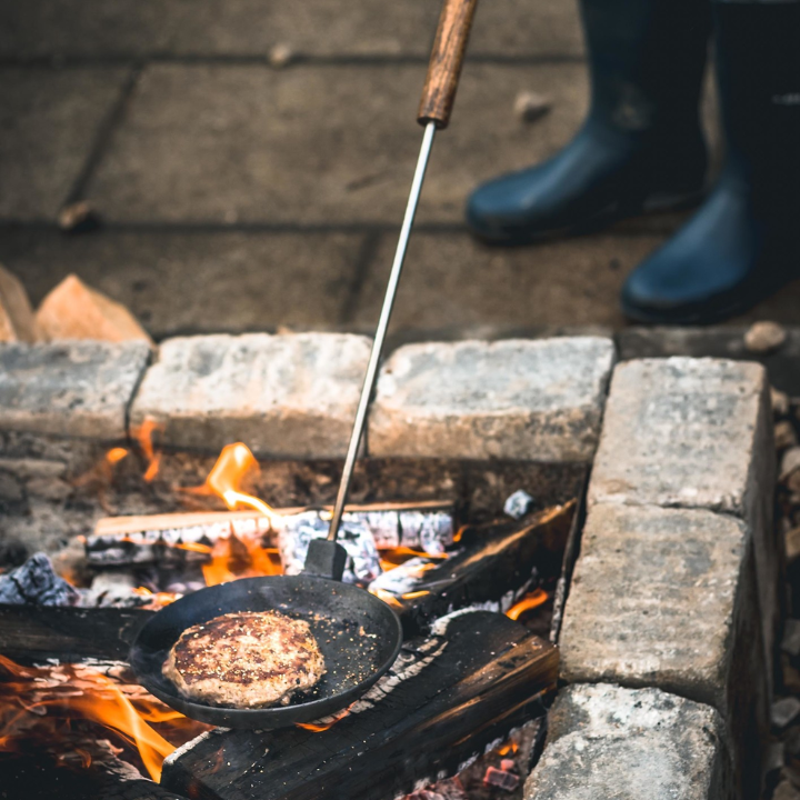 Een zwarte, ronde kookpan met houten handvat en metalen stang, voorzien van een kleine zilveren bel aan de bovenkant. Geschikt voor het koken van omeletten en andere gerechten op de kookplaat.