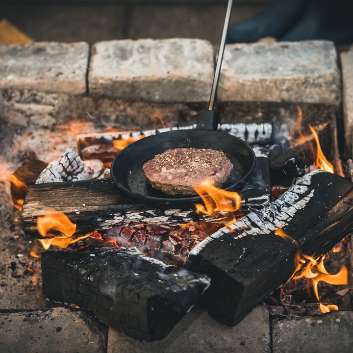 Zwarte kookpan met houten handvat en metalen steel, geschikt voor koken op kampvuur of grill. Gebruik voor het bereiden van eenvoudige maaltijden in de natuur.