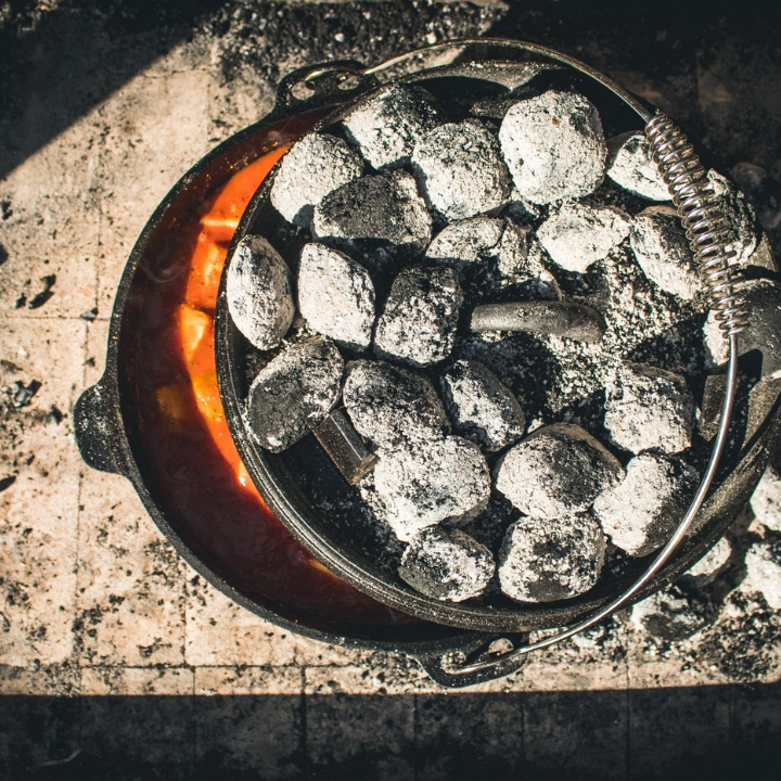 Zwarte, glanzende kookpan van gietijzer met afdekking, voorzien van handgrepen en een stijlvolle, gegraveerde tekst. Ideaal voor outdoor koken en hoge hittebestendigheid.