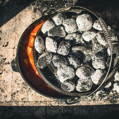 Zwarte, glanzende kookpan van gietijzer met afdekking, voorzien van handgrepen en een stijlvolle, gegraveerde tekst. Ideaal voor outdoor koken en hoge hittebestendigheid.