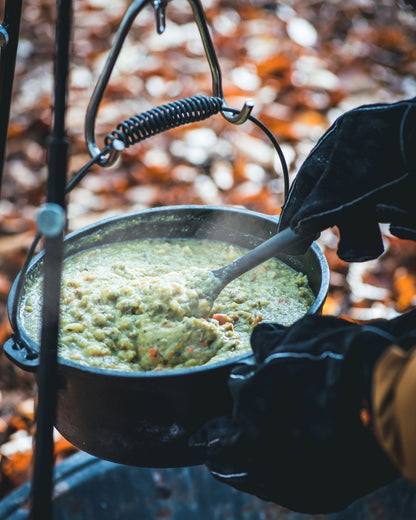 Zwarte kookpan van gietijzer met een robuuste, gebogen metalen handgreep en een springveer. Geschikt voor kampvuur en outdoor koken, met een gladde binnenkant en handige oorschelpen.