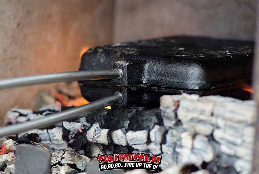 Een zwarte, vierkante roosterplaat met een geëmbosseerd kruis, verbonden aan een lichtbruine, geribbelde houten handgreep via een metalen stang, geschikt voor barbecueën en buitenkoken.